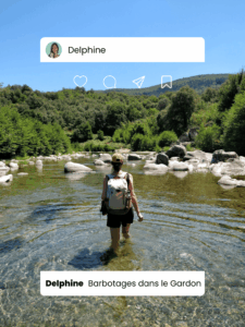 Femme de dos marchant dans une rivière peu profonde entourée de galets et de végétation dense, sac à dos beige sur les épaules, sous un ciel bleu clair.